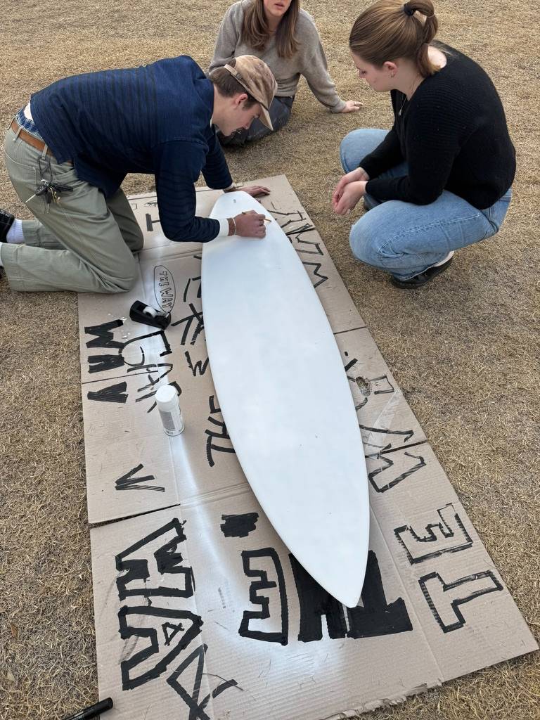 Three people kneeling around a blank surfboard on cardboard covered in lettering drafts